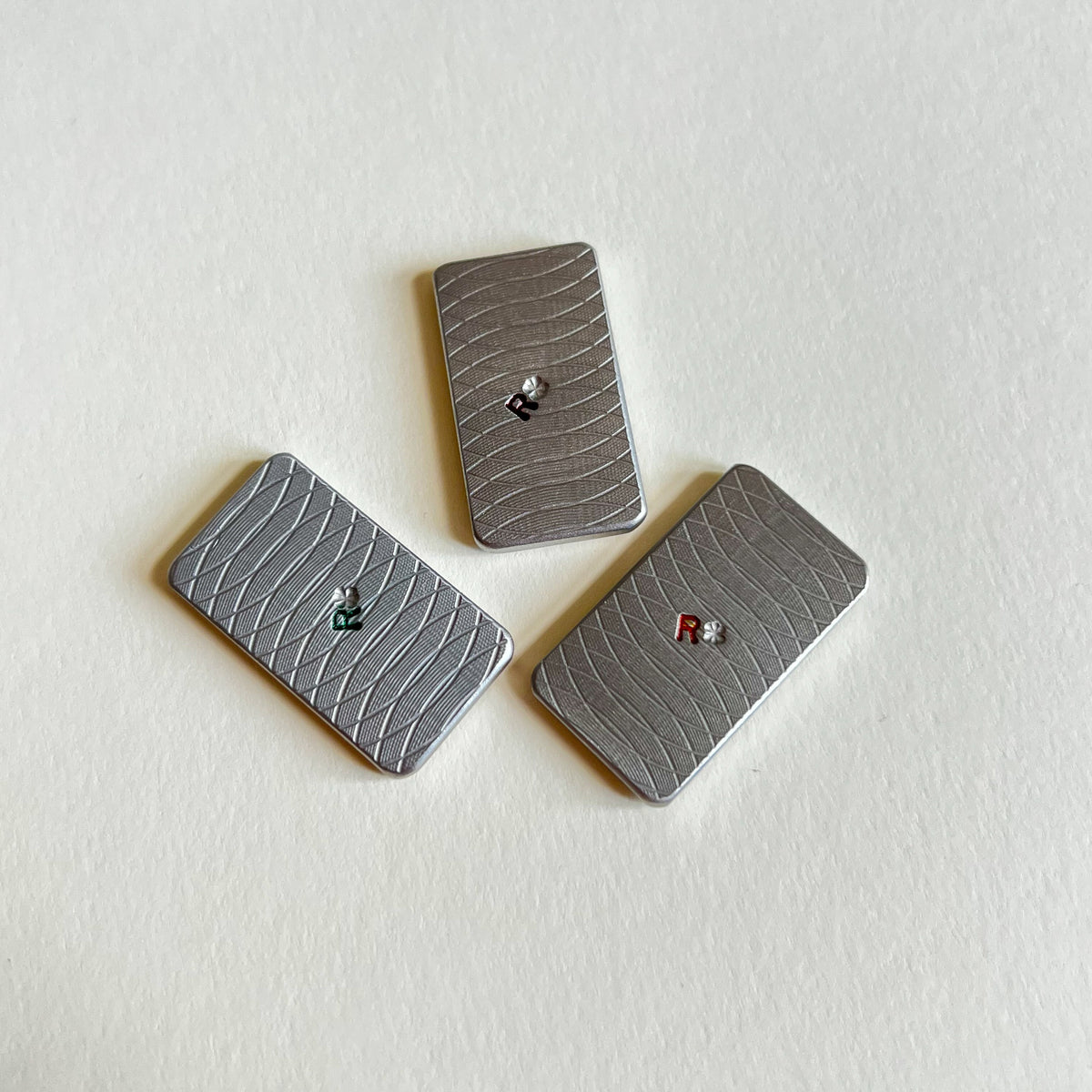 Three stainless steel metal golf ball markers with a textured diamond pattern, arranged diagonally on a white background. 