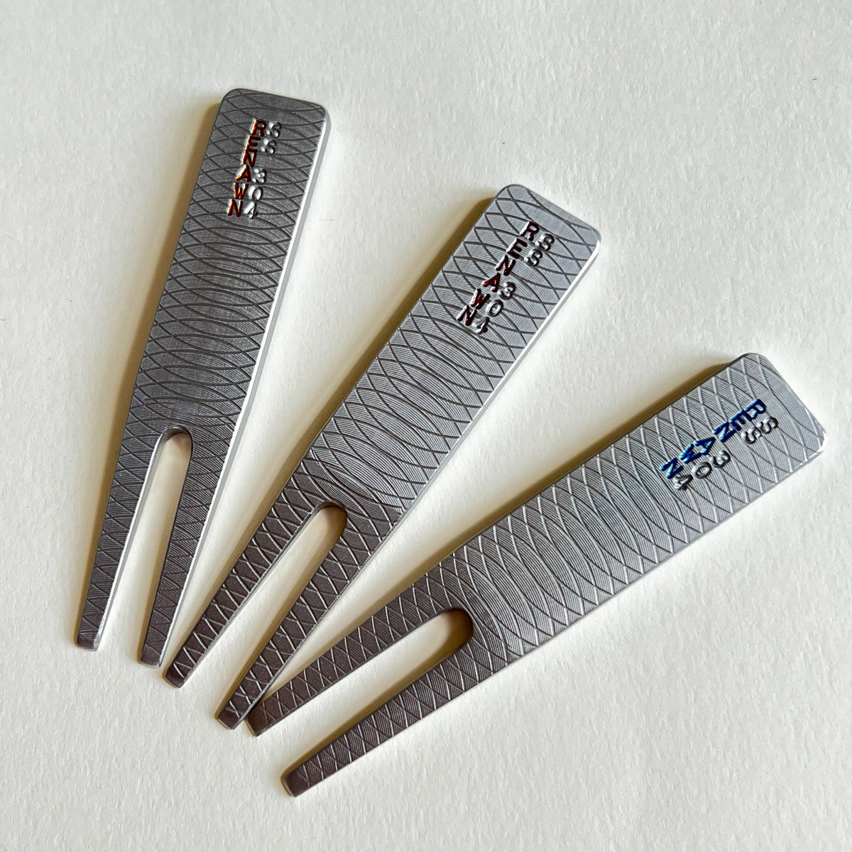Three stainless steelmetal golf divottools with a textured diamond pattern, arranged diagonally on a white background. 